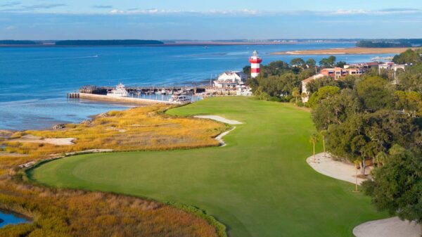 Aerial view of the 18th hole at Harbour Town Golf Links, featuring the closing fairway along the Calibogue Sound with the iconic red-and-white lighthouse in the background.