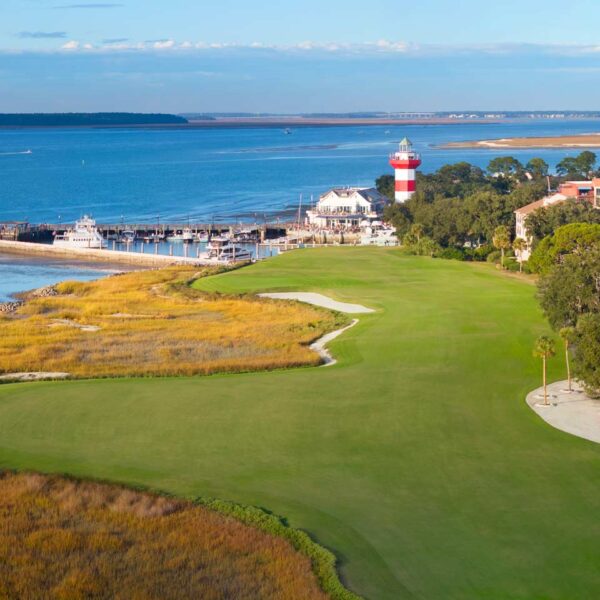 Aerial view of the 18th hole at Harbour Town Golf Links, featuring the closing fairway along the Calibogue Sound with the iconic red-and-white lighthouse in the background.