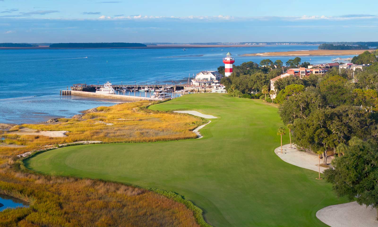 Aerial view of the 18th hole at Harbour Town Golf Links, featuring the closing fairway along the Calibogue Sound with the iconic red-and-white lighthouse in the background.
