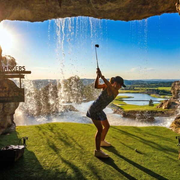 Ozarks golf at Cliffhangers Lion’s Den, featuring a golfer teeing off from a cave through a waterfall toward a scenic green in Branson, Missouri